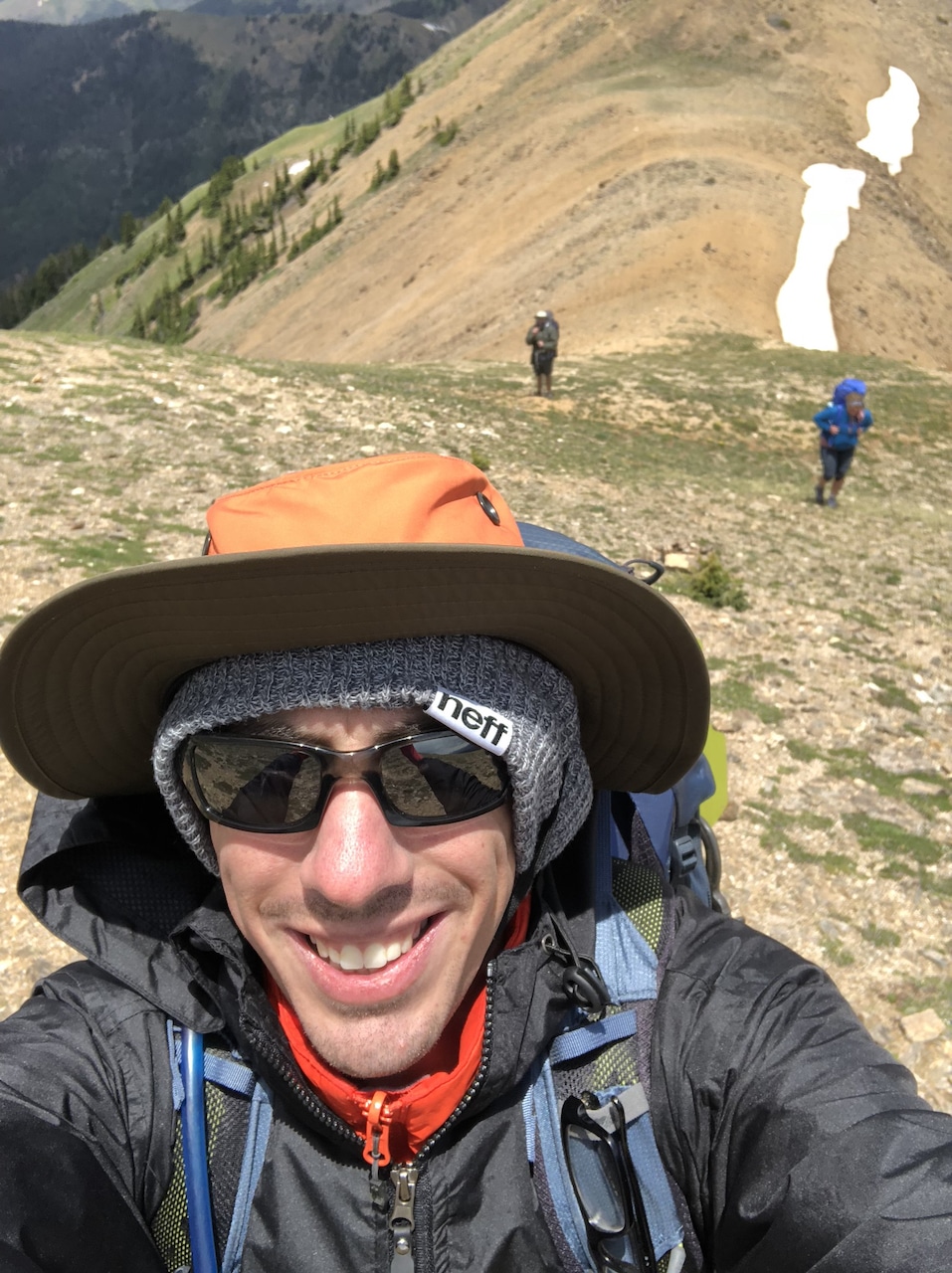 Selfie of Matt wearing a jacket and hat at the top of a partially snow-covered mountain with two friends in the background.